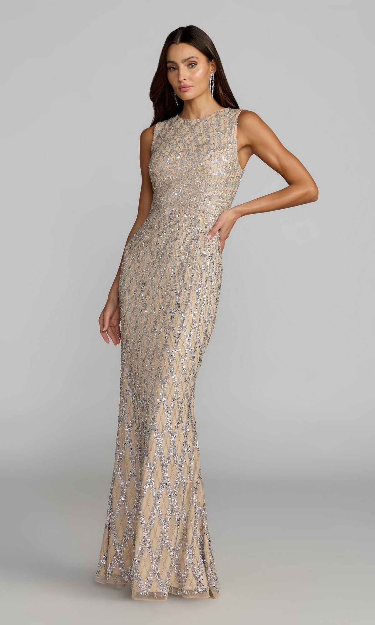 A woman poses with one hand on her hip against a plain gray background, wearing the Long Formal Dress: Mac Duggal 6249 in Nude Ombre, a sleeveless, floor-length gown embellished with silver geometric sequin patterns.